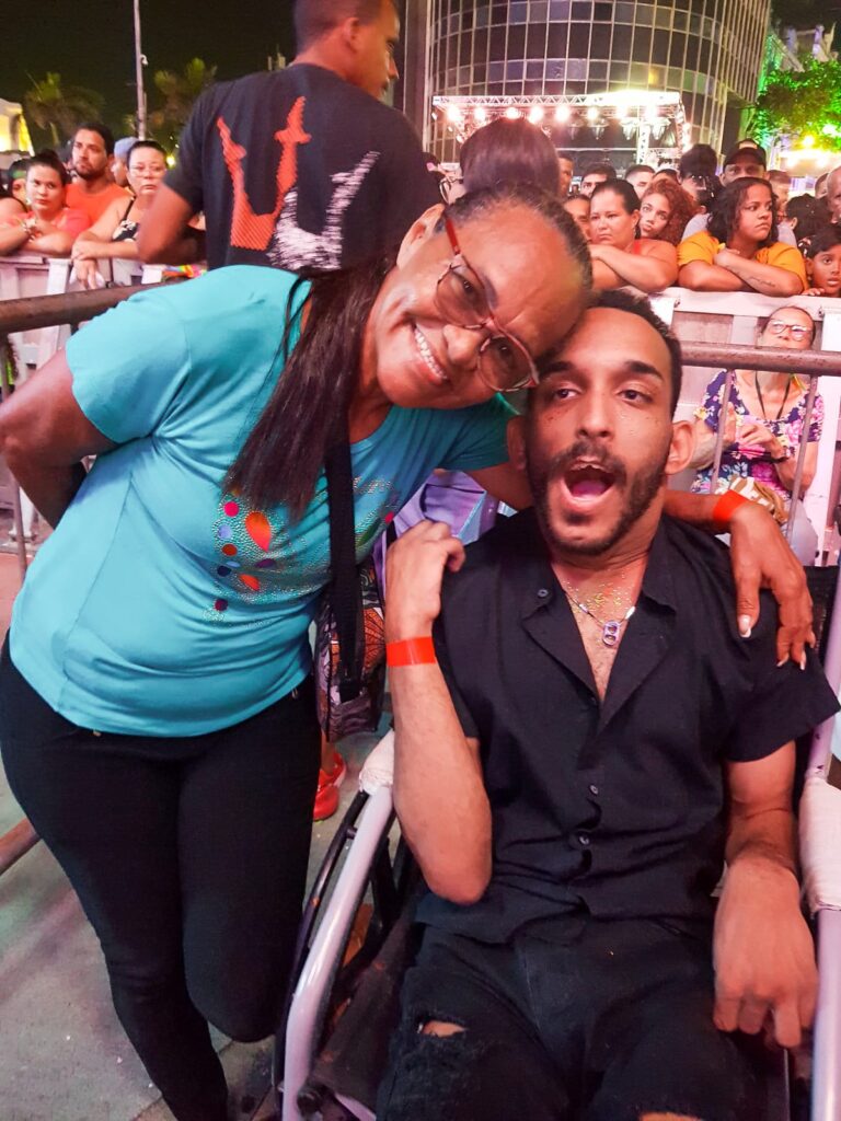 Fotografia de Vânia, uma mulher negra, com cabelo com rabo de cabelo, uma blusa azul e calça jeans e está abraçando Jonatas um homem negro, com cabelo curtinho, usa barba, está blusa e caça preta e está sentando na cadeiras de rodas.