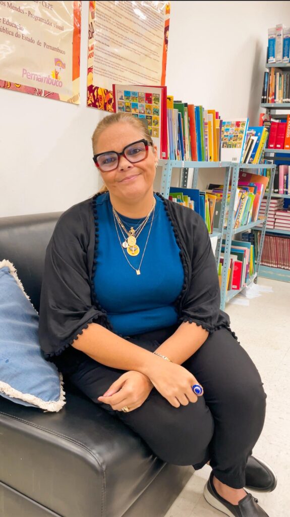 fotografia de uma mulher branca, com cabelos loiros presos, usando óculos grandes, vestindo uma blusa azul e um casaco preto por cima. Ela está sentada em um sofá preto e usa colares e anéis dourados. Ao fundo, há estantes com livros e cartazes nas paredes.