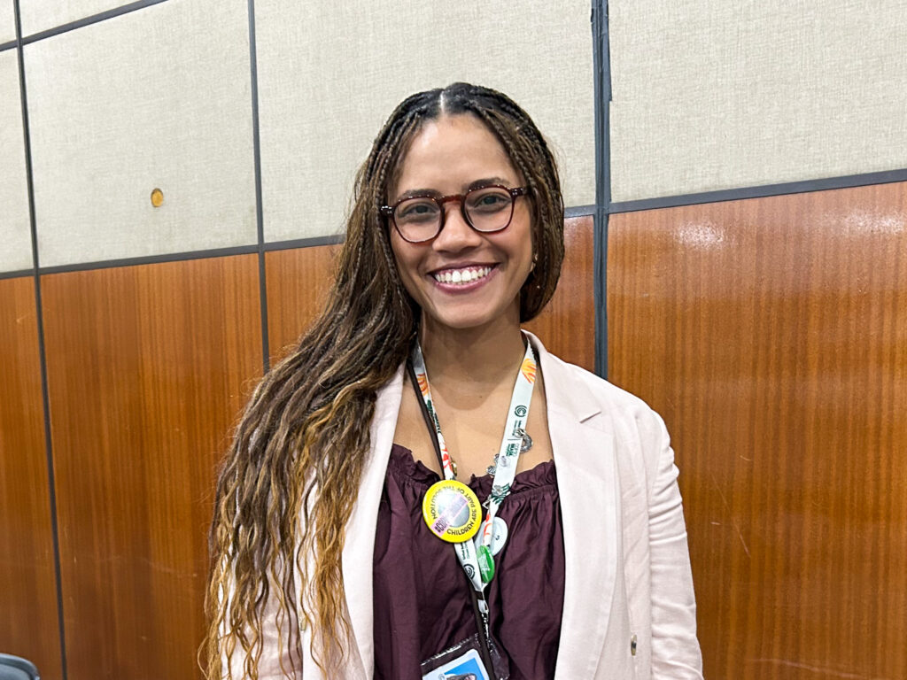 Uma mulher jovem, de cabelos longos com tranças finas, sorri para a câmera. Ela usa óculos de armação arredondada, blazer claro e blusa vinho. No pescoço, veste um crachá com vários adesivos. Ao fundo, há uma parede de painéis acústicos em tons de bege e madeira.