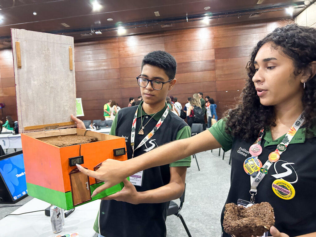 Dois adolescentes apresentam uma maquete colorida de uma casa. O jovem à esquerda segura a estrutura aberta, enquanto a jovem à direita aponta para os detalhes frontais da maquete. Ambos usam uniforme escolar escuro com crachás e adesivos pendurados. No fundo, há outras mesas, pessoas circulando e painéis de madeira escuros na parede.