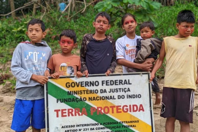Seis crianças indígenas estão em pé. Elas têm expressão séria. Suas peles estão marcadas com pintura vermelha no rosto. Elas seguram uma placa retangular e traz o seguinte texto em destaque: “Governo Federal — Ministério da Justiça — Fundação Nacional do Índio. Terra Protegida. Acesso interditado a pessoas estranhas. ”O fundo mostra vegetação.