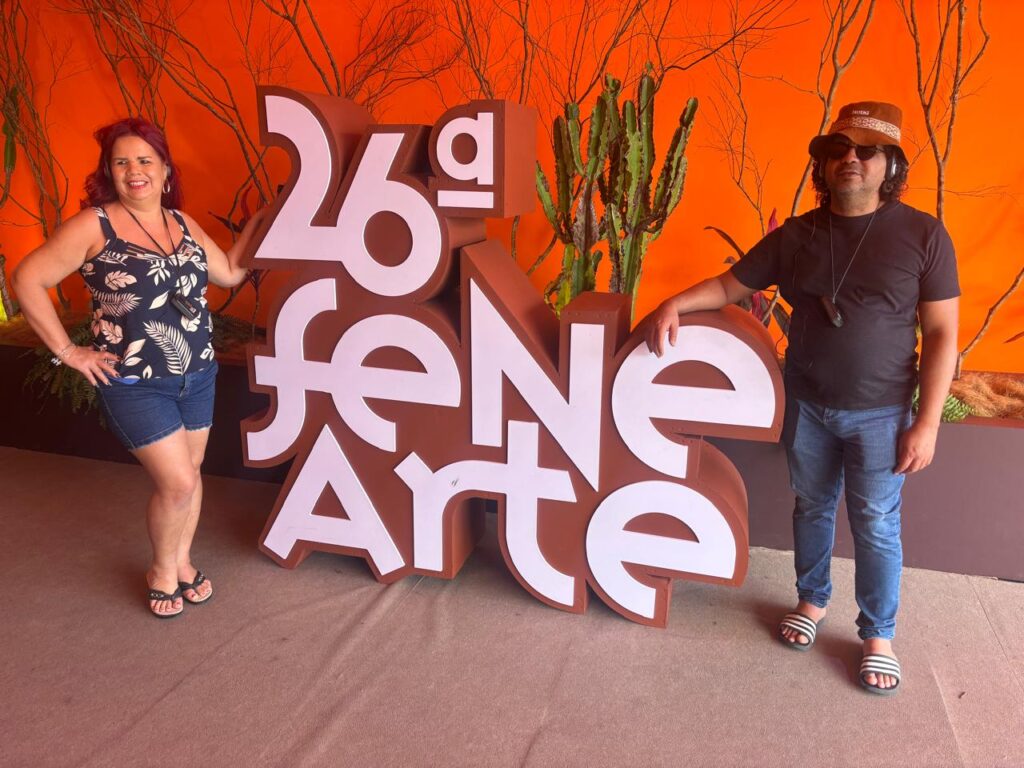 Foto de duas pessoas posando ao lado da estrutura cenográfica da "26ª Fenearte", em letras grandes brancas e marrons. À esquerda, uma mulher de cabelos avermelhados usa blusa estampada de fundo preto, short jeans e sandálias pretas. À direita, um homem usa chapéu marrom, óculos escuros, camiseta preta, calça jeans e sandálias. Ambos sorriem para a foto e apoiam as mãos na estrutura da Fenearte. Ao fundo, um painel laranja, galhos secos e cactos.