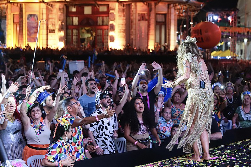 Fotografia de uma plateia de pessoas com deficiência voltada para o palco onde Elba Ramalho se apresenta, com um vestido dourado.