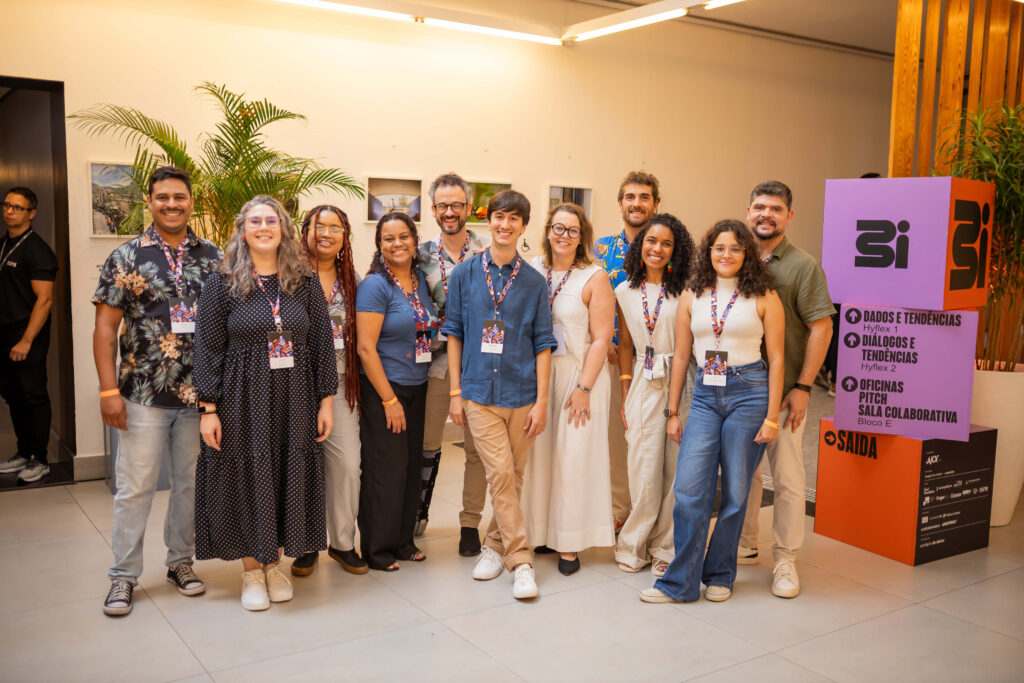 Doze pessoas posa sorridente no festival 3i. Todos usam crachás e roupas casuais. Ao fundo, parede clara com fotos e planta. À direita, sinalização colorida indica salas do evento. 