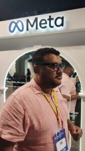 imagem robson homem negro, cabelo pretos curtos, usando oculos preto, camisa rosa e ao fundo painel bege com logotipo meta.