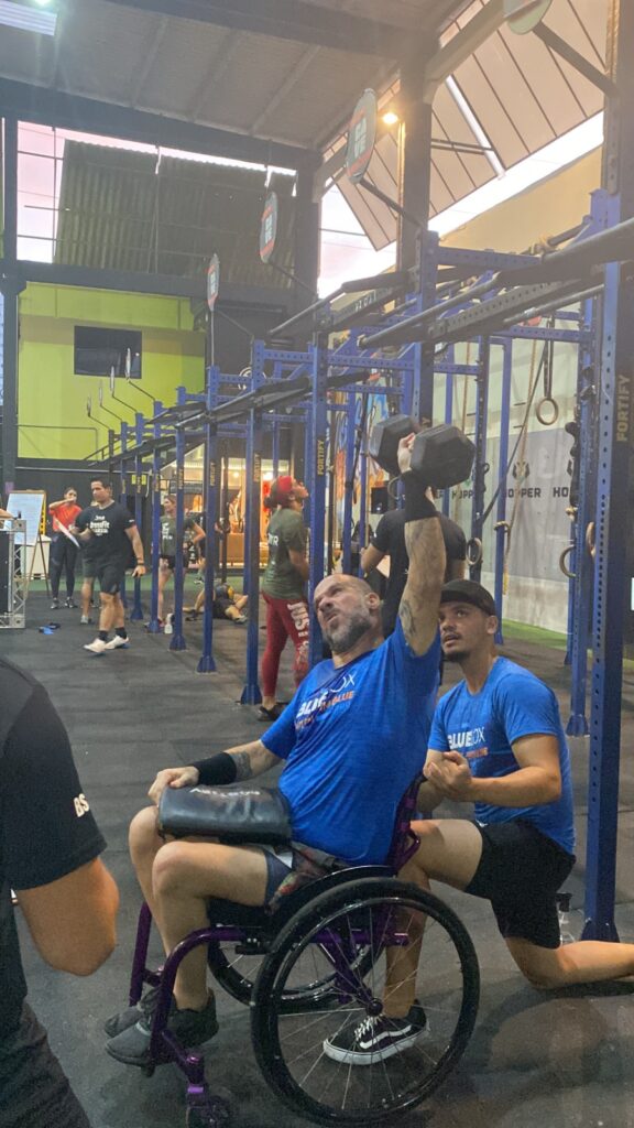 Pedro sentado em uma cadeira de rodas, ergue um haltere preto acima da cabeça com o braço direito. Ele usa camiseta azul e munhequeira preta. Ao lado dele, um homem blusa azul. Ao fundo, há outras pessoas treinando.