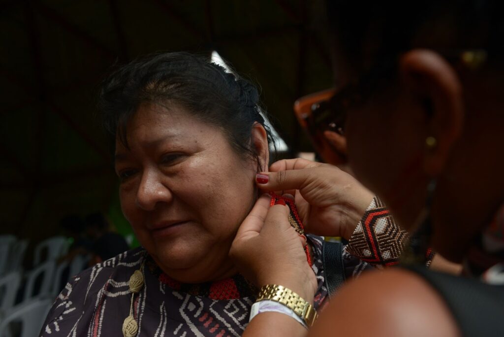 Fotografia de Joenia, uma mulher indígena de cabelo preto preso. Ela usa uma blusa estampada e uma pessoa coloca um brinco na orelha as mãos.