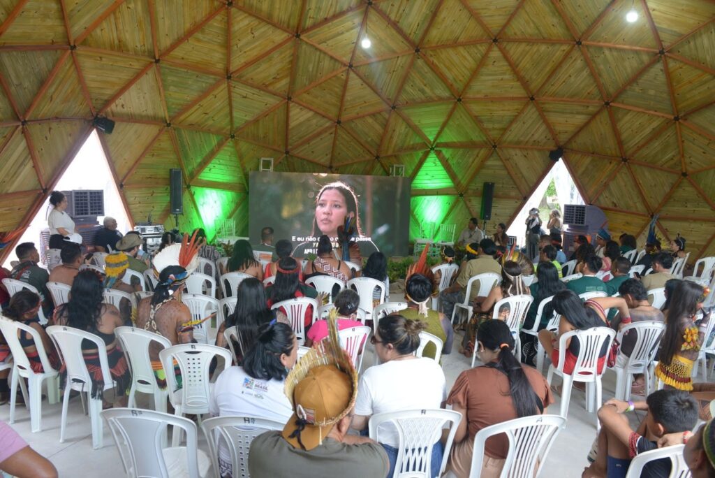 Fotografia de uma sala arredondada, com estrutura de palha e madeira. Várias pessoas estão sentadas, olhando para um telão que exibe a imagem de uma mulher indígena.