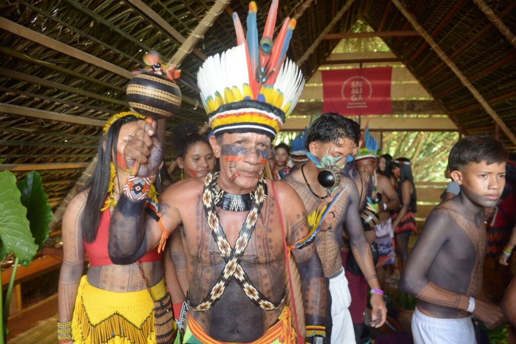 Fotografia de um grupo de indígenas com pinturas corporais e cocares, alinhados em fileira.