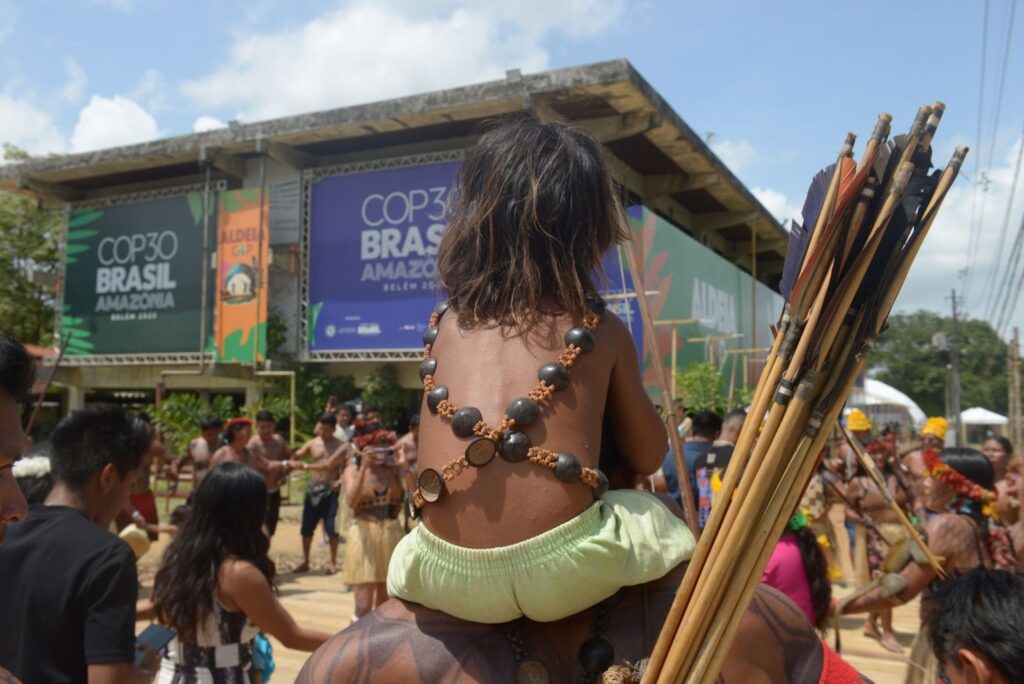 Fotografia de uma criança de costas, apoiada nos ombros de uma pessoa com várias pinturas corporais e lanças. Ao fundo, aparece um prédio com um banner da COP30 Brasil.
