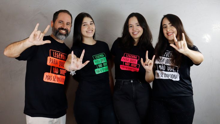 Quatro pessoas posam lado a lado em frente a uma parede clara. Da esquerda para a direita: um homem de barba grisalha e três mulheres de cabelos escuros. Todos vestem camisetas pretas com a frase “Legenda para quem não ouve, mas se emociona!” escrita em diferentes cores (Laranja, verde, rosa e branca). Eles fazem com as mãos o sinal de “eu te amo” em Libras e sorriem para a câmera