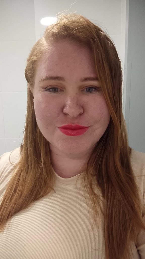 Fotografia em plano médio de uma mulher de pele clara e cabelos ruivos longos e lisos. Ela está de frente, olhando diretamente para a câmera, com batom vermelho nos lábios. Usa uma blusa bege de gola alta e tecido canelado. Ao fundo paredes brancas, com iluminação vinda do topo.